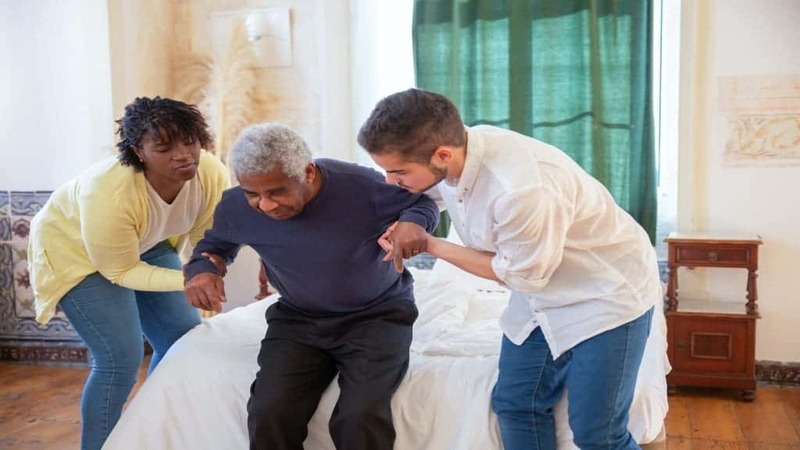Two caregivers help an elderly person stand from a bed in a bright room with wooden floors and green curtains.